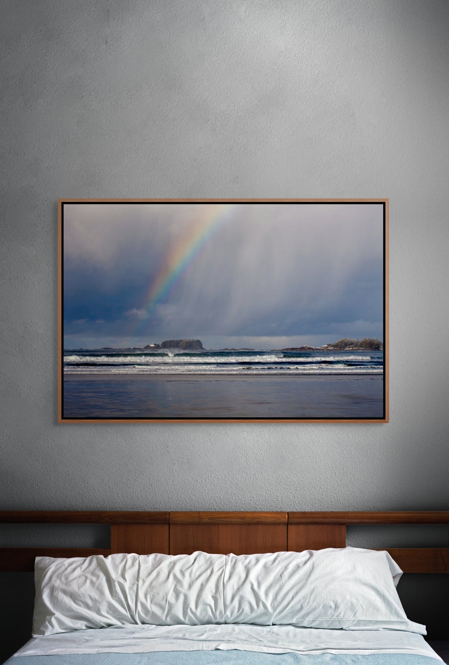 Framed artwork of a rainbow over the surf of the Pacific Ocean on a gray wall above a bed. Cedar float frame.