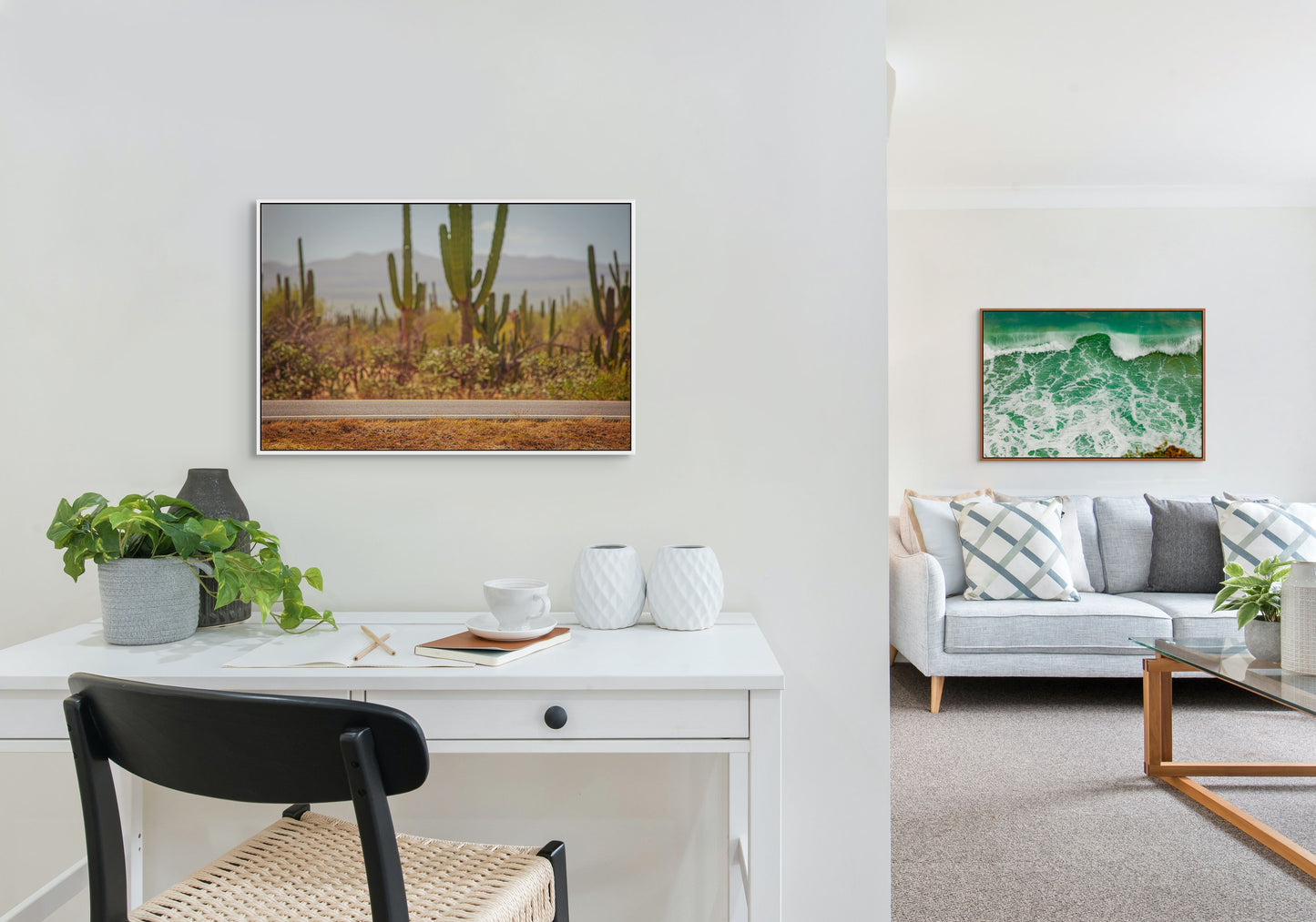 White framed artwork of a desert road on a wall above a white desk in a modern living room with a cedar framed artwork of a wave above a couch in the right side of the image in the background.