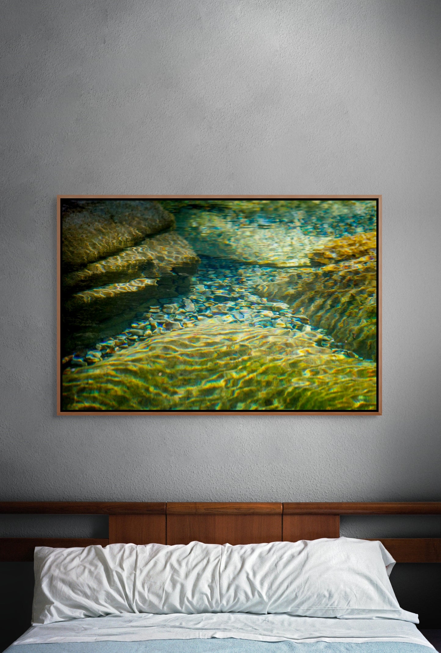 Framed artwork of clear water flowing over rocks with sunlight filtering through above a bed with white pillows and blue bedding.