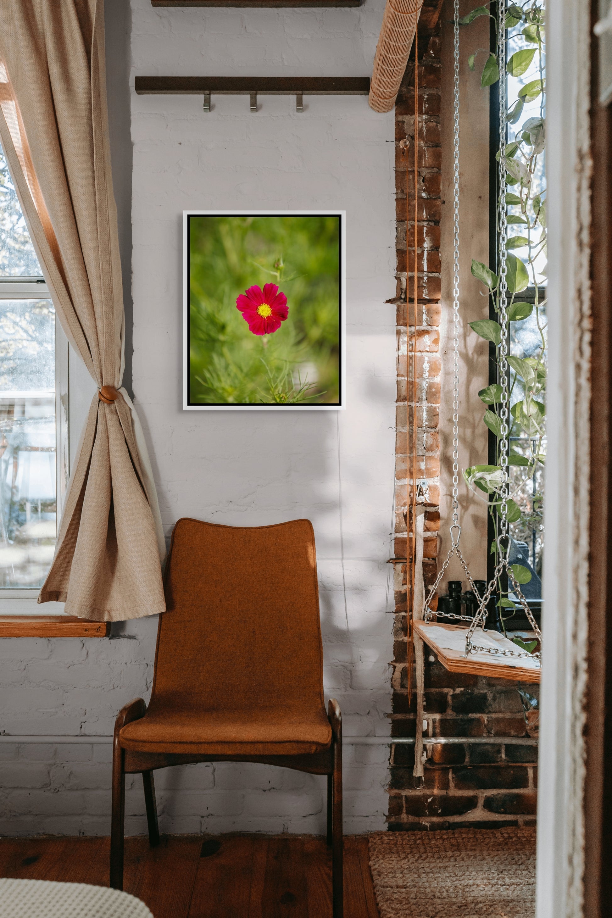 Room interior with a framed picture of a red flower on a white wall, an orange chair, and a window with curtains.
