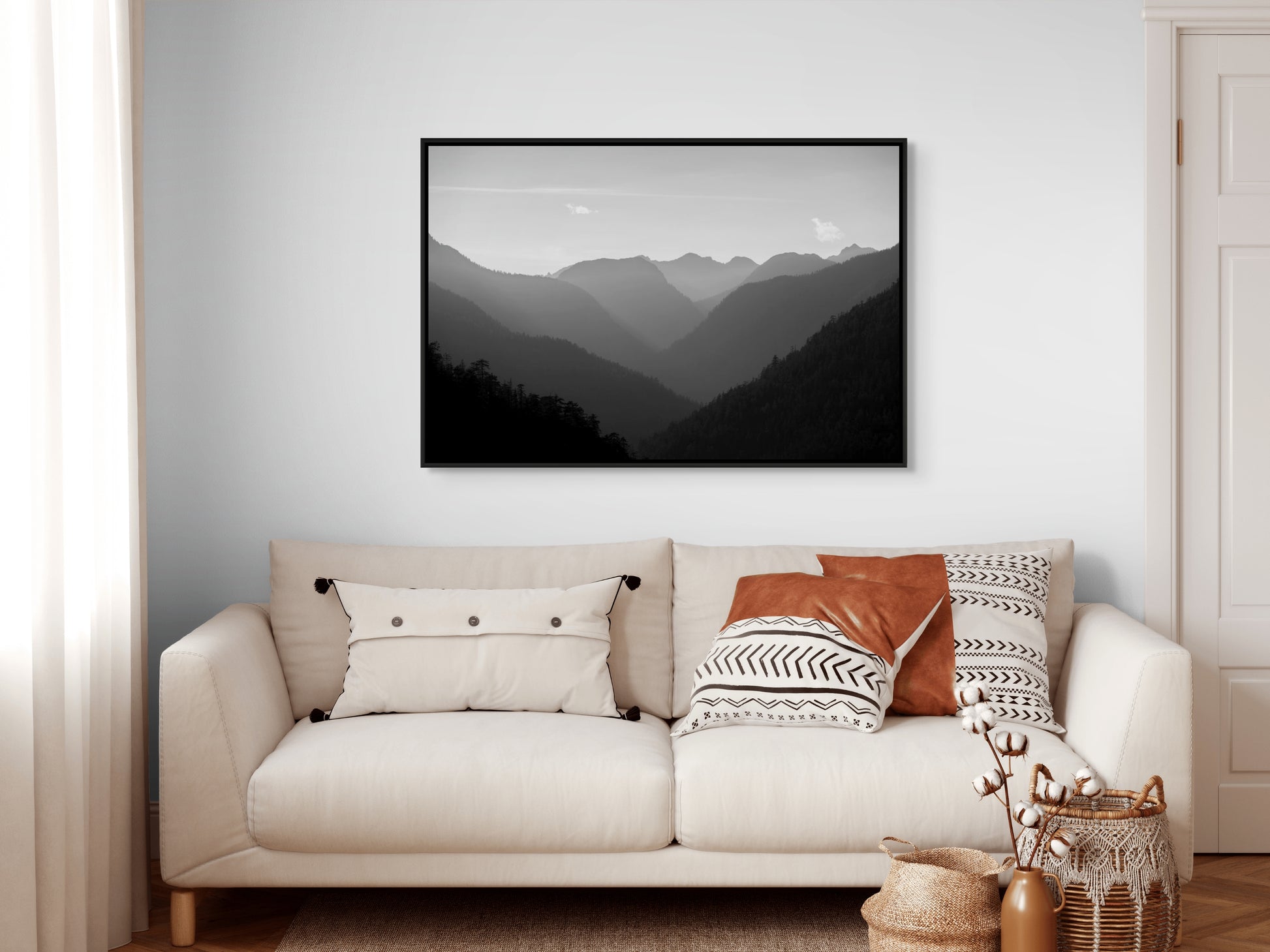 Beige sofa with decorative pillows and a black framed mountain landscape painting on a white wall.