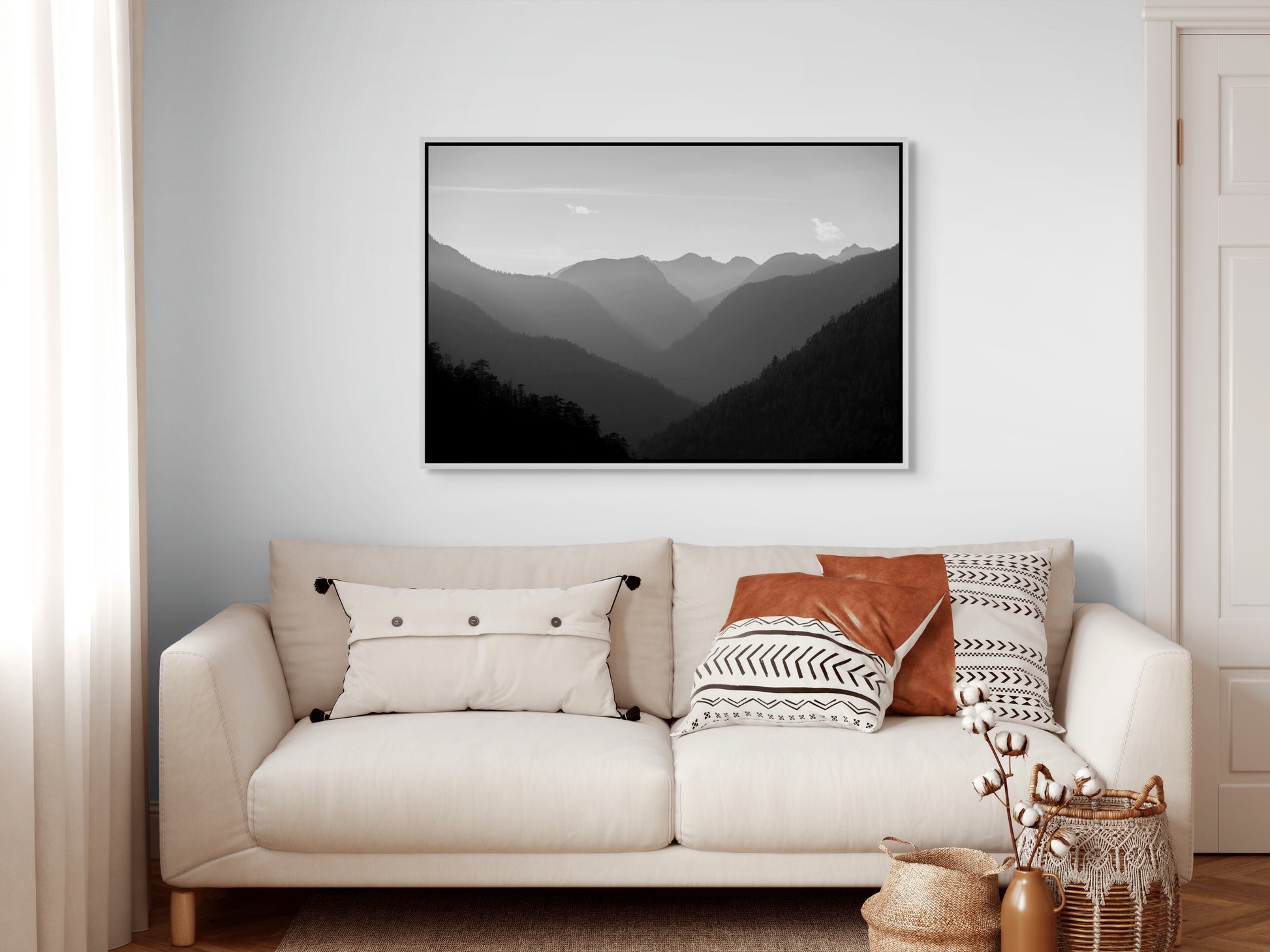 Beige sofa with decorative pillows and a white framed mountain landscape painting on a white wall.
