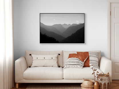 Beige sofa with decorative pillows and a white framed mountain landscape painting on a white wall.