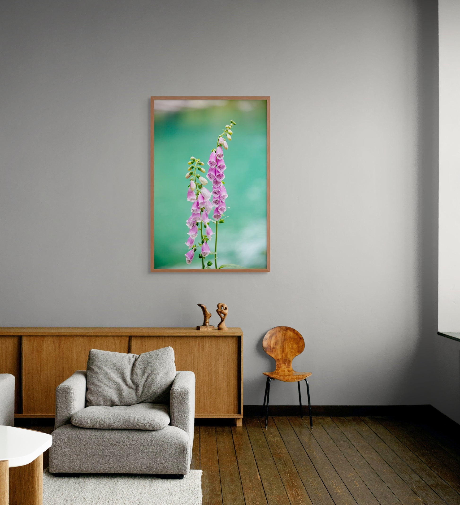 Cedar framed artwork of pink flowers on a gray wall above a gray armchair and wooden cabinet.