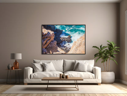 Modern living room with a white sofa, coffee table, tropical plant, and a black framed art print of a turquoise ocean meeting the rocky shore.