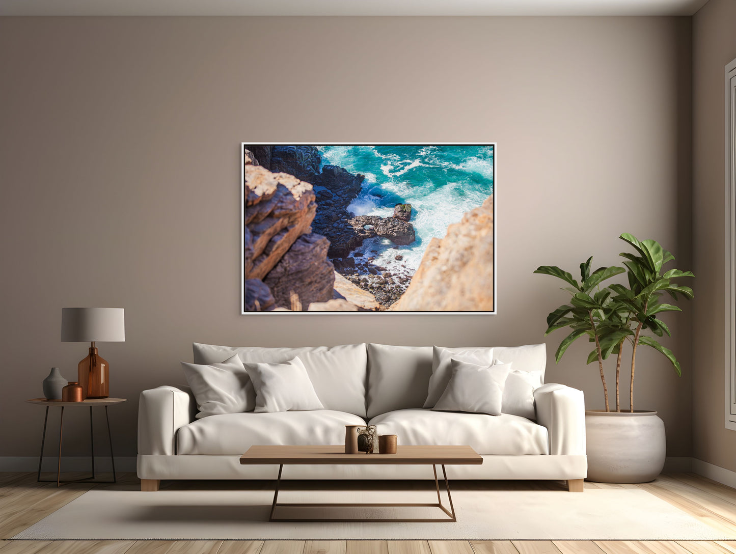 Modern living room with a white sofa, coffee table, tropical plant, and a white framed art print of a turquoise ocean meeting the rocky shore.