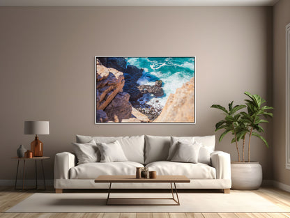 Modern living room with a white sofa, coffee table, tropical plant, and a white framed art print of a turquoise ocean meeting the rocky shore.