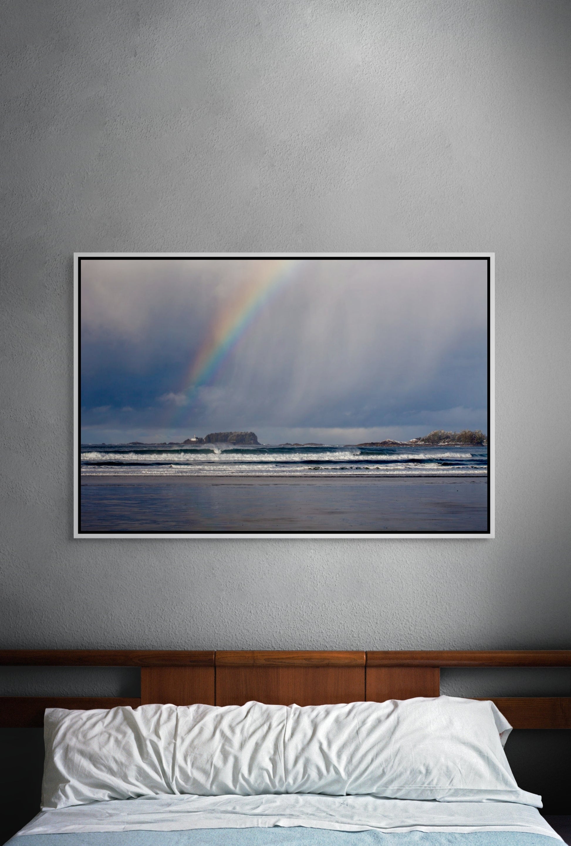 Framed artwork of a rainbow over the surf of the Pacific Ocean on a gray wall above a bed. White float frame.