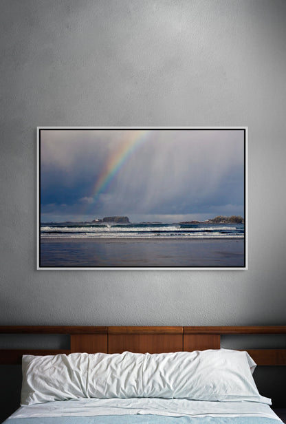 Framed artwork of a rainbow over the surf of the Pacific Ocean on a gray wall above a bed. White float frame.