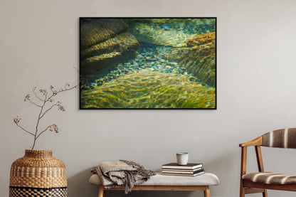 Living room with a large framed picture of clear water flowing over rocks with sunlight filtering through on the wall, a woven vase, and a chair. Black float frame.