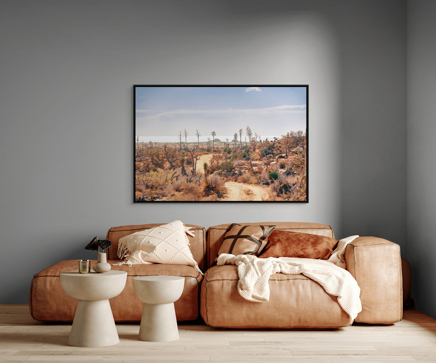 Living room with a brown leather sofa, two white stools, and a large black framed photo print on a gray wall depicting a desert landscape.