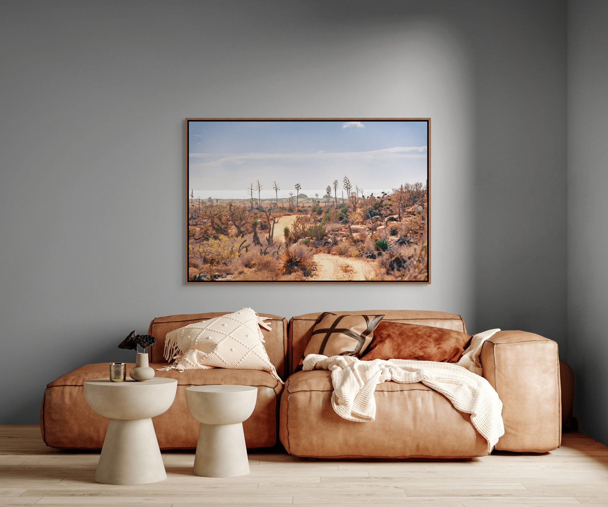 Living room with a brown leather sofa, two white stools, and a large cedar framed photo print on a gray wall depicting a desert landscape.