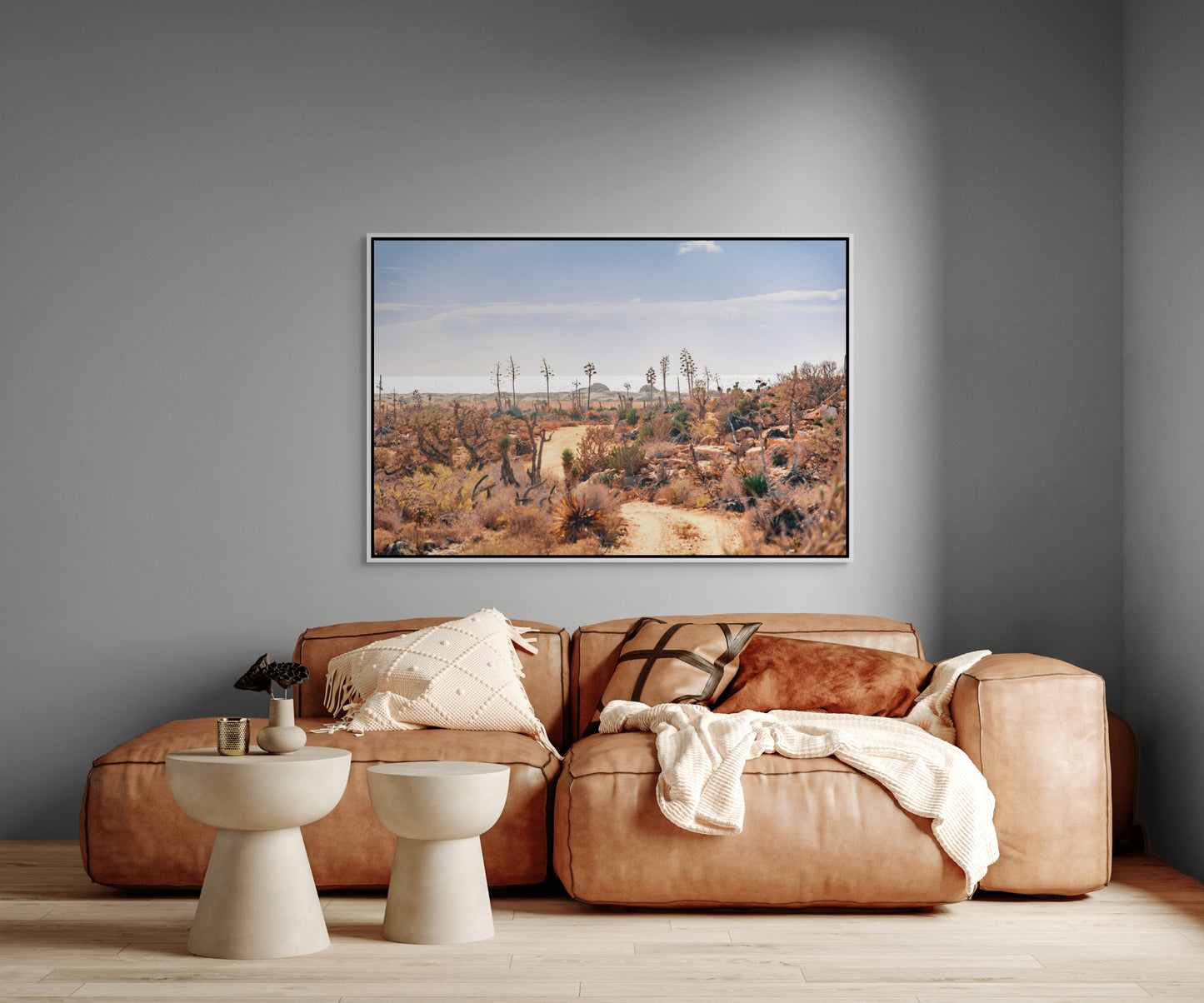 Living room with a brown leather sofa, two white stools, and a large white framed photo print on a gray wall depicting a desert landscape.